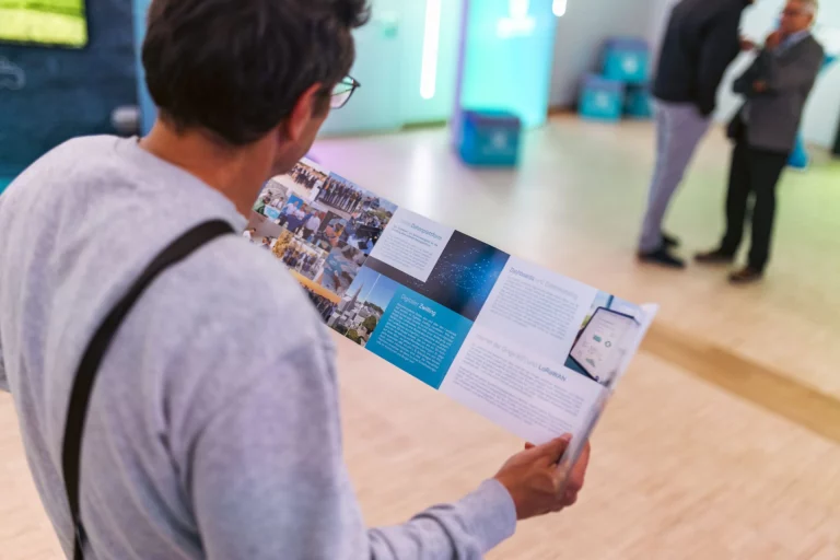 Mann betrachtet Informationsfaltblatt, im Hintergrund Personen im Gespräch