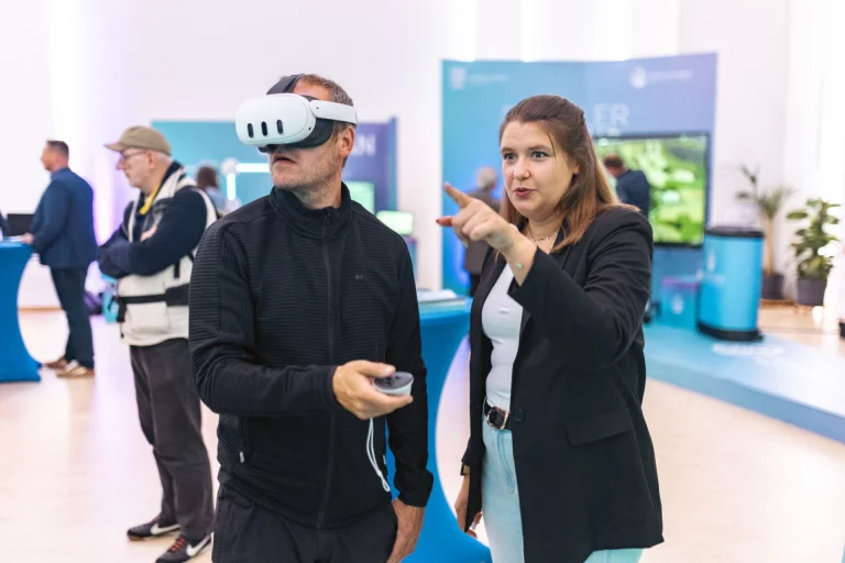 Mann mit VR-Brille und Controller neben Frau, die auf einen Bildschirm im Ausstellungsraum zeigt