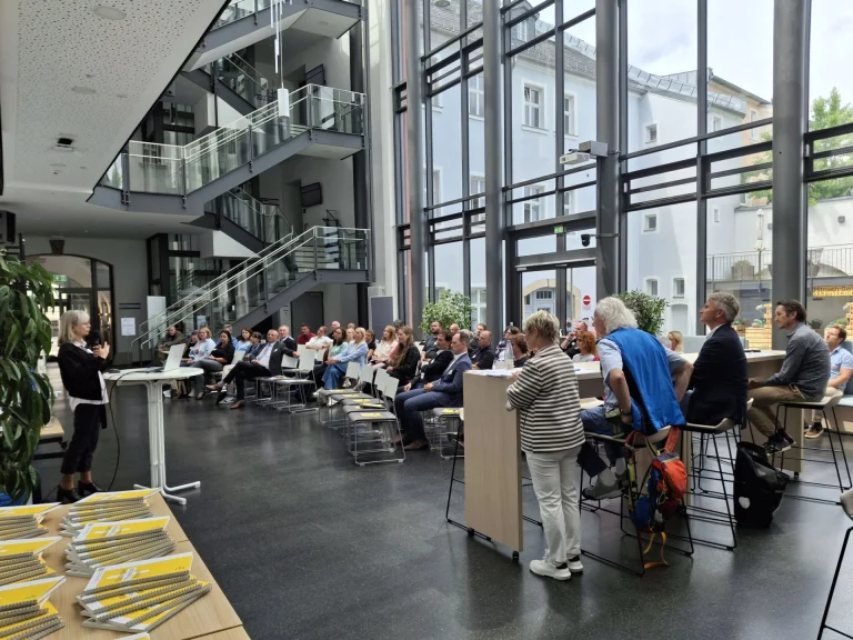 Vortrag im Foyer eines Gebäudes mit Treppenhaus vor sitzendem und stehendem Publikum
