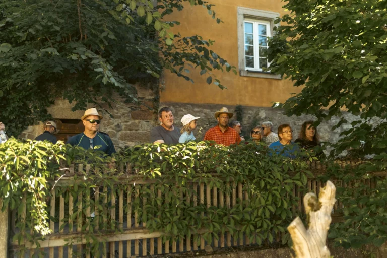Zuschauergruppe beobachtet von einer Gartenterrasse aus hinter Gebüsch das Geschehen