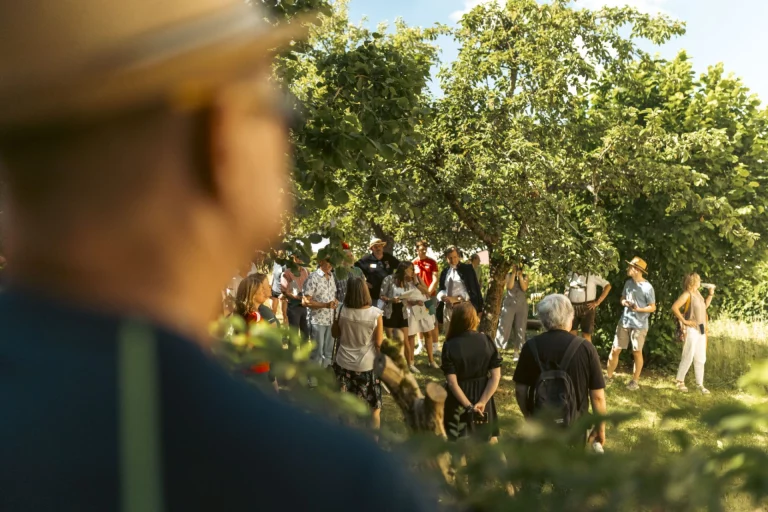 Besuchergruppe steht im Streuobstgarten unter Bäumen, im Vordergrund unscharf eine Person mit Hut