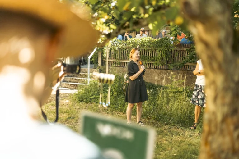Frau mit Mikrofon spricht im Garten vor Publikum und Fotograf mit Teleobjektiv