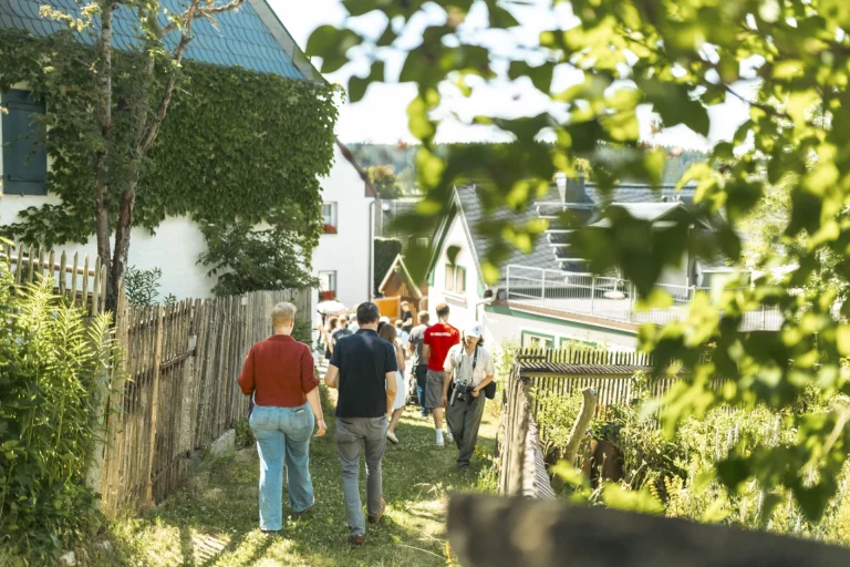 Gruppe von Personen geht auf schmalem Weg zwischen Gärten durch das Dorf