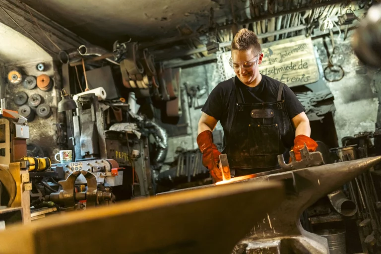 Person in Schürze und Schutzbrille bearbeitet glühendes Metall an einem Amboss in der Schmiede
