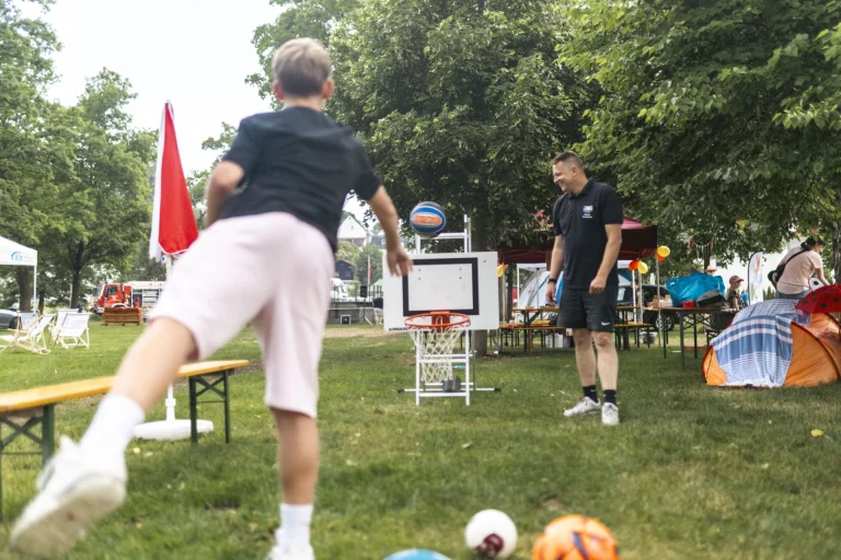 Junge wirft Basketball auf mobilen Korb, im Hintergrund lachender Mann und Zelte auf der Festwiese