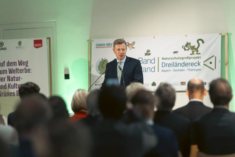 Mann am Rednerpult vor Plakaten zum Naturschutzgroßprojekt Grünes Band Dreiländereck, Publikum im Vordergrund