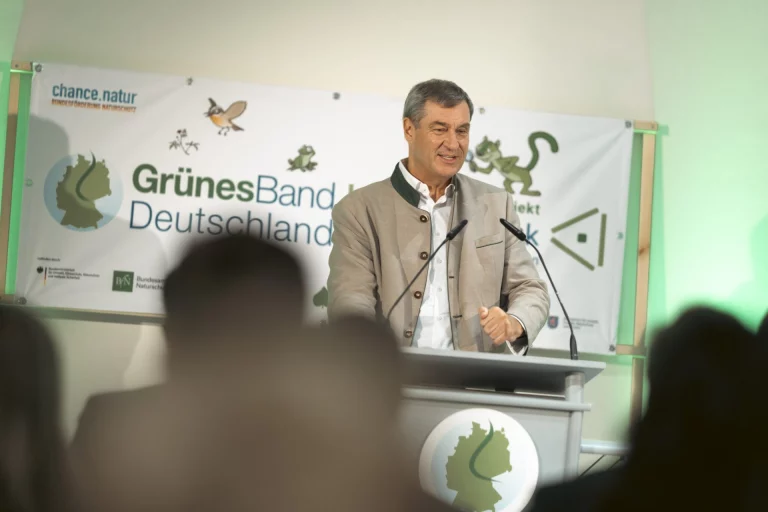 Mann im Trachtenjanker spricht am Rednerpult vor Banner „Grünes Band Deutschland“, im Vordergrund Zuhörende