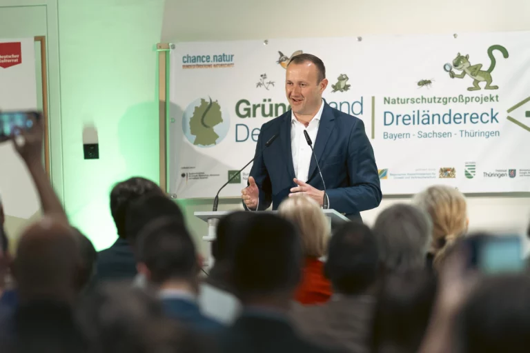 Mann im Anzug spricht am Rednerpult vor Publikum, dahinter Banner „Grünes Band Dreiländereck“