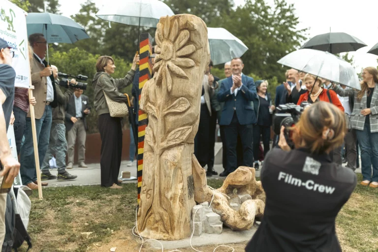 Enthüllung einer Holzskulptur bei der Eröffnungsfeier, umringt von Gästen unter Regenschirmen und einer Film-Crew