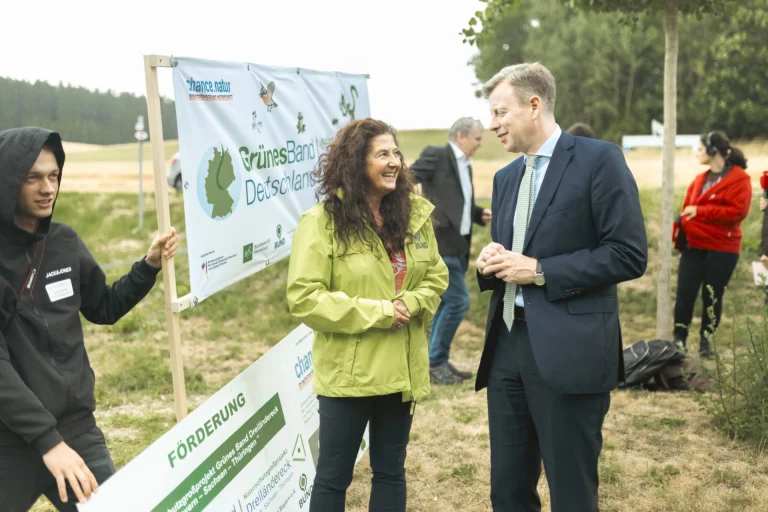 Frau in grüner Jacke und Mann im Anzug im Gespräch neben Banner „Grünes Band Deutschland“