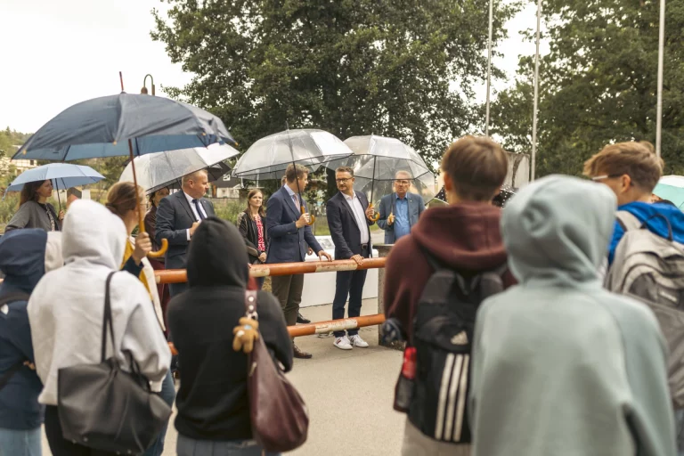 Gruppe mit Regenschirmen hört Männern in Mänteln am historischen Grenzzaun in Mödlareuth zu