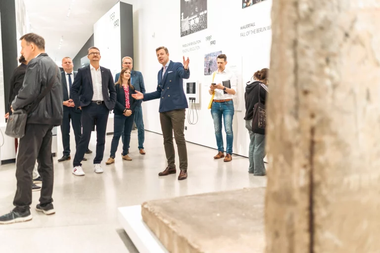 Gruppe von Personen in einer Ausstellung vor Schautafeln zur deutschen Teilung, im Vordergrund ein Mauersegment