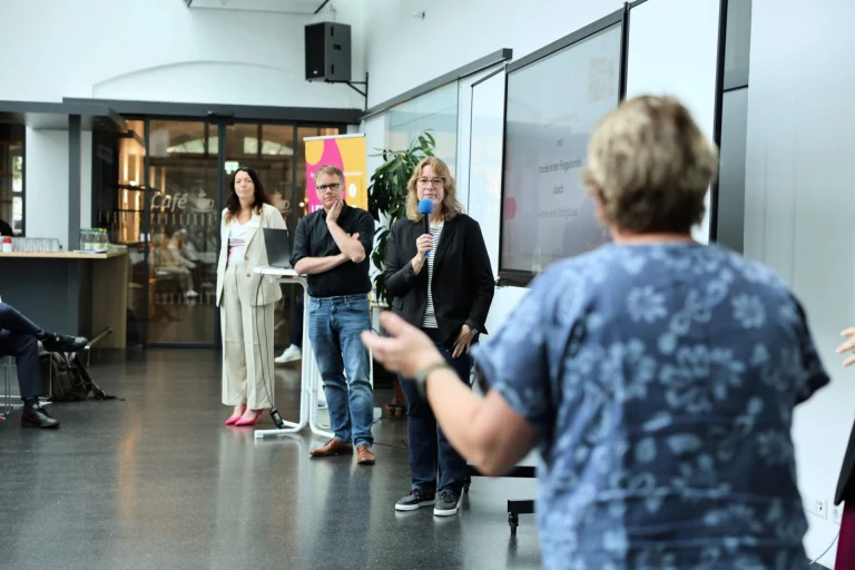 Frau mit Mikrofon spricht zu einer Gruppe im Foyer, im Hintergrund zwei weitere Personen am Stehtisch