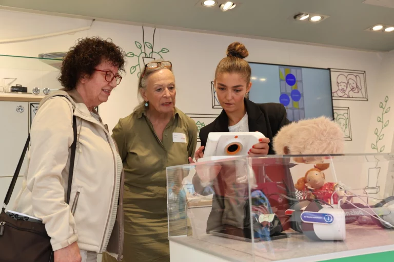 Zwei Besucherinnen und eine Mitarbeiterin betrachten Pflegehilfsmittel an einem Infostand