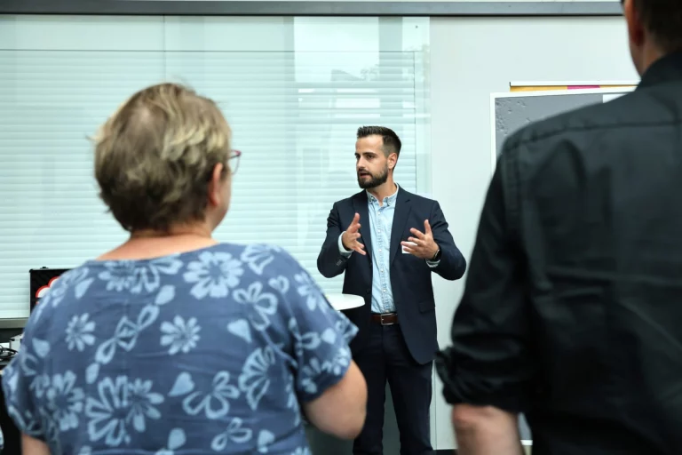 Mann in Sakko spricht gestikulierend zu zwei Personen im Besprechungsraum