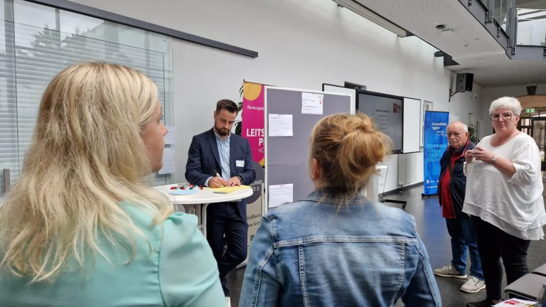 Gespräch zwischen Teilnehmenden vor Infotafeln und Beratungsstand beim Runden Tisch Pflege