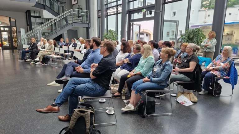 Mehrere Personen sitzen aufgereiht im Foyer und verfolgen eine Veranstaltung