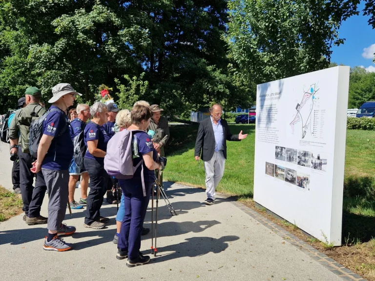 Wandergruppe mit Wanderstöcken und Rucksäcken steht vor einer Infotafel, ein Mann erläutert sie