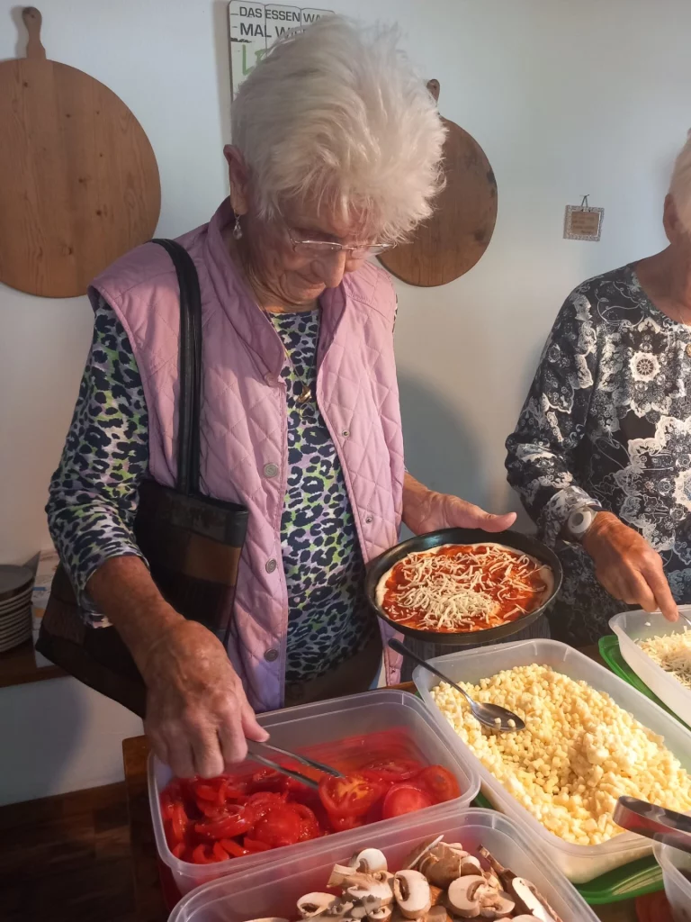 Ältere Frau belegt Pizzateig mit Tomaten und Käse an einem Buffet