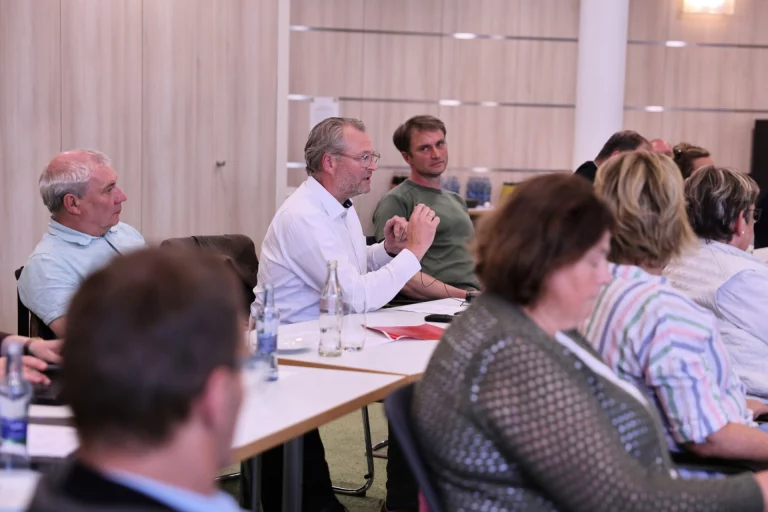 Teilnehmende des Waldsymposiums sitzen an Tischen, ein Mann spricht mit erhobener Hand
