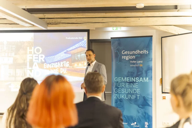 Mann hält Vortrag neben Roll-up Gesundheitsregion plus Hofer Land, Zuhörende im Vordergrund
