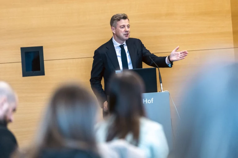 Mann im Anzug spricht am Rednerpult mit Aufschrift Hof vor Publikum