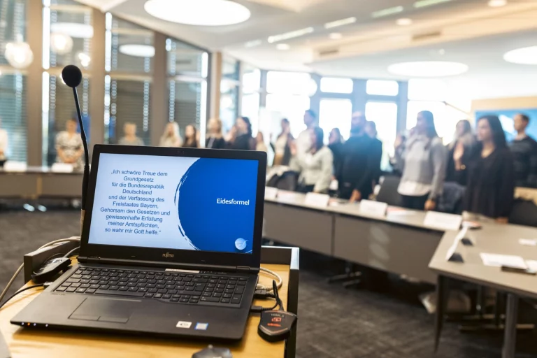 Laptop mit Eidesformel auf dem Bildschirm, im Hintergrund stehende Gruppe mit erhobener Hand