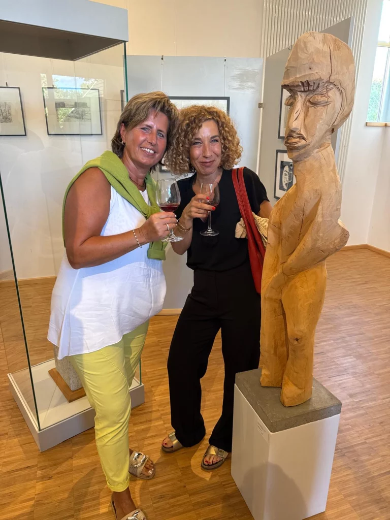Zwei Frauen mit Weinglas neben einer Holzskulptur in der Ausstellung