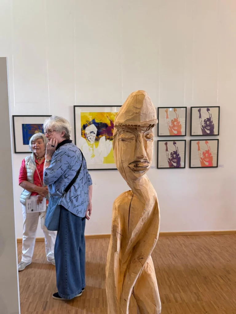 Zwei Personen betrachten eine Holzskulptur und gerahmte Kunstwerke in einer Ausstellung