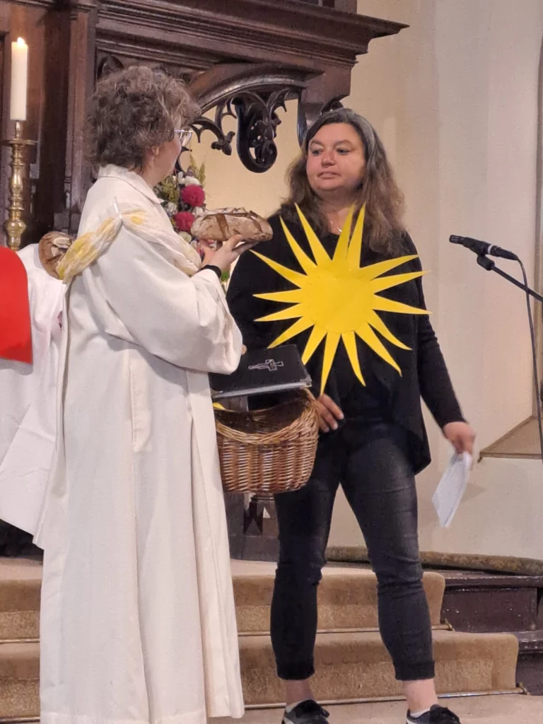Pfarrerin überreicht Brot an Frau mit großer gelber Sonnensymbol-Applikation am Altar