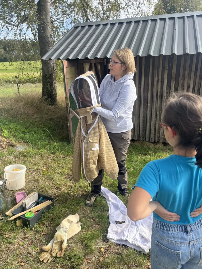 Frau erklärt Imker-Schutzkleidung im Freien vor einem Bienenhaus, Mädchen schaut zu